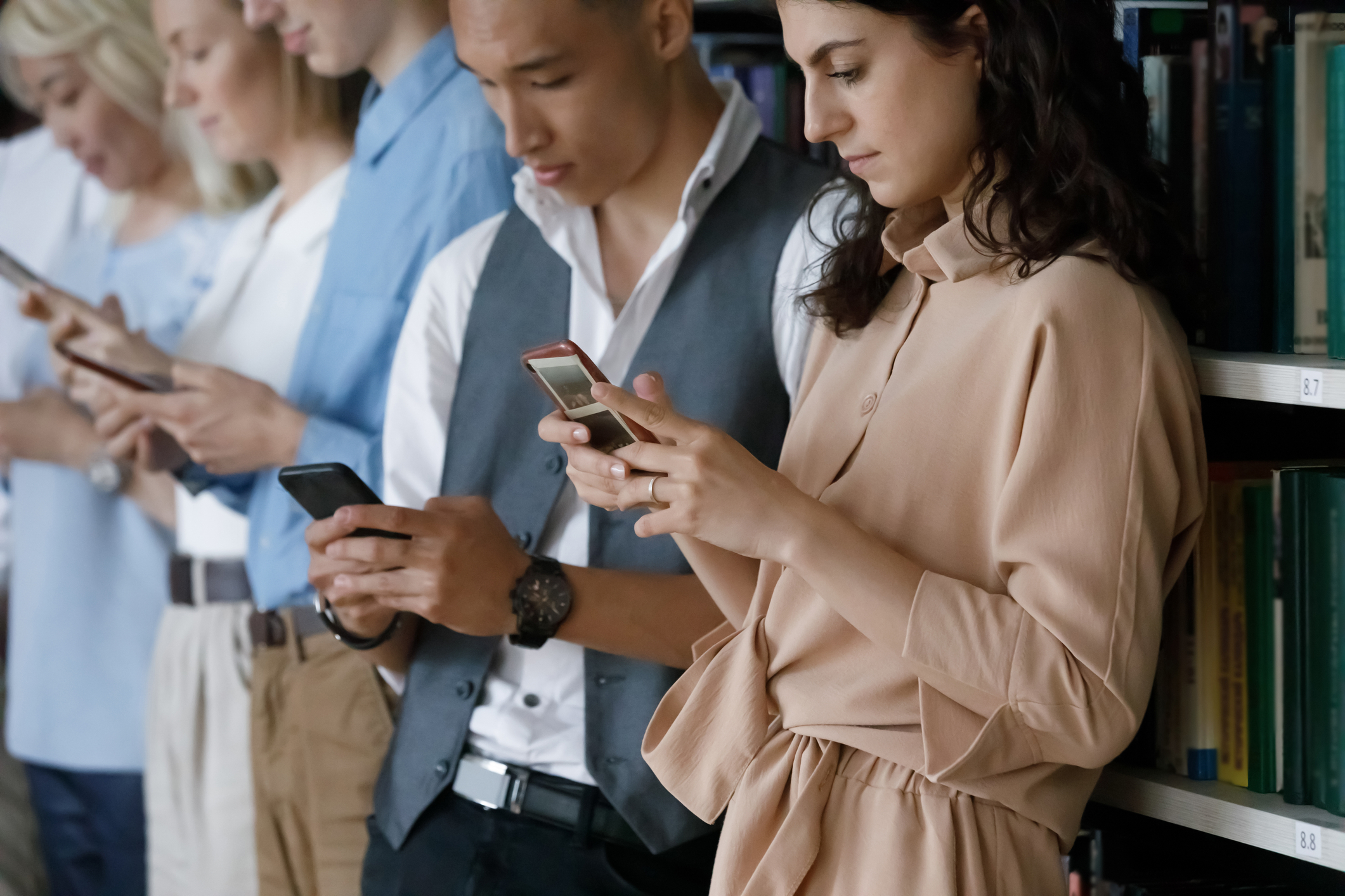 Line of people standing near book shelves, all looking at cell phones.