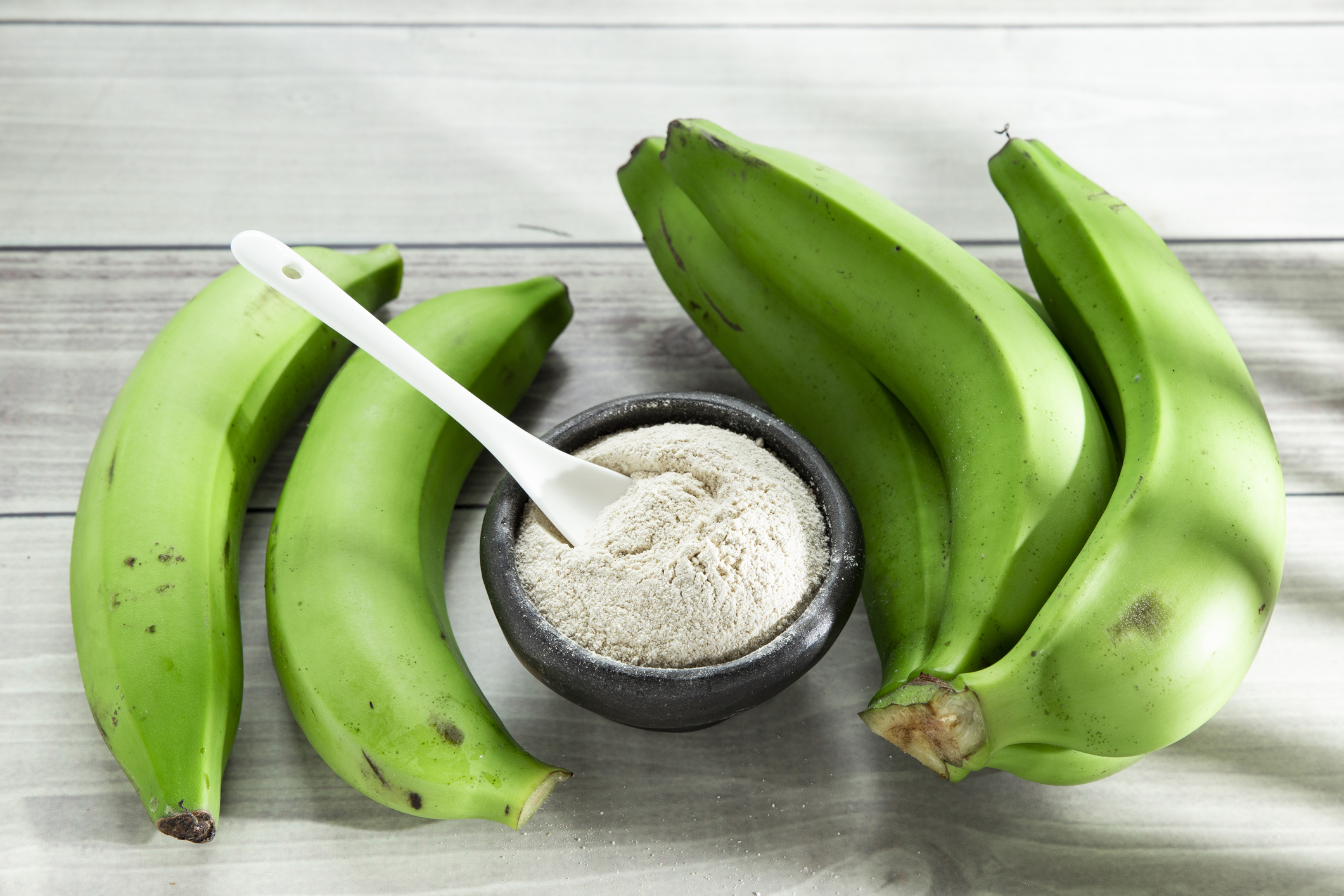 A small bowl of green banana powder with a spoon in it surrounded by green bananas,