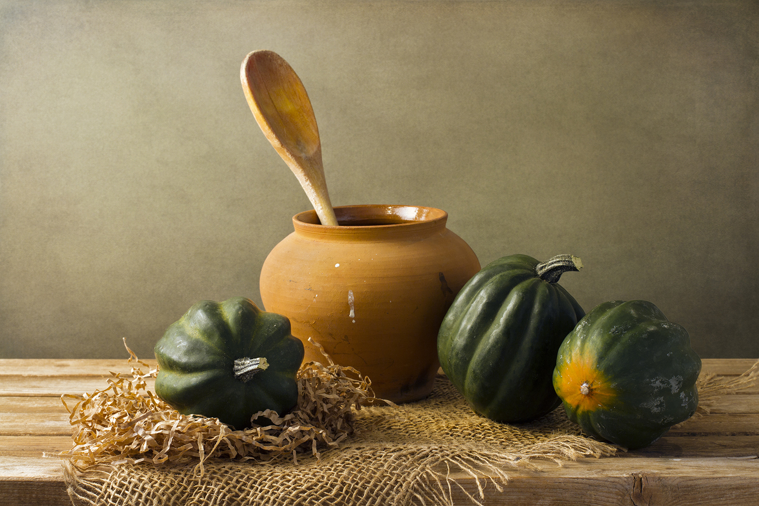 Whole acorn squash on a rustic table next to an earthen vase with a wooden spoon i it.