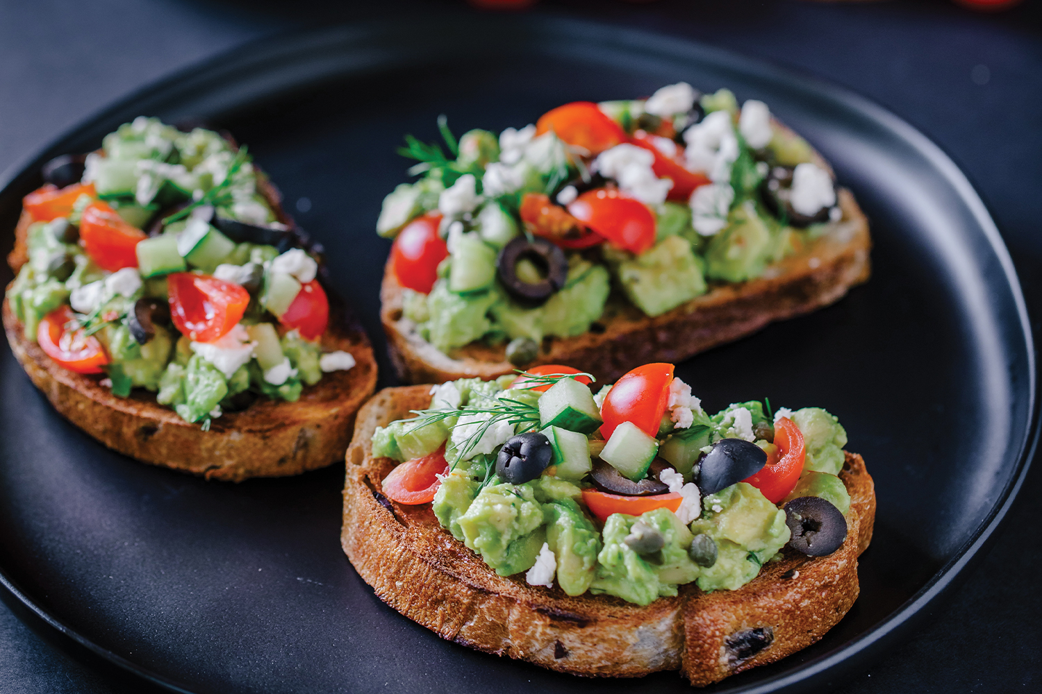 Greek Avocado Toast on a black plate with a black background.