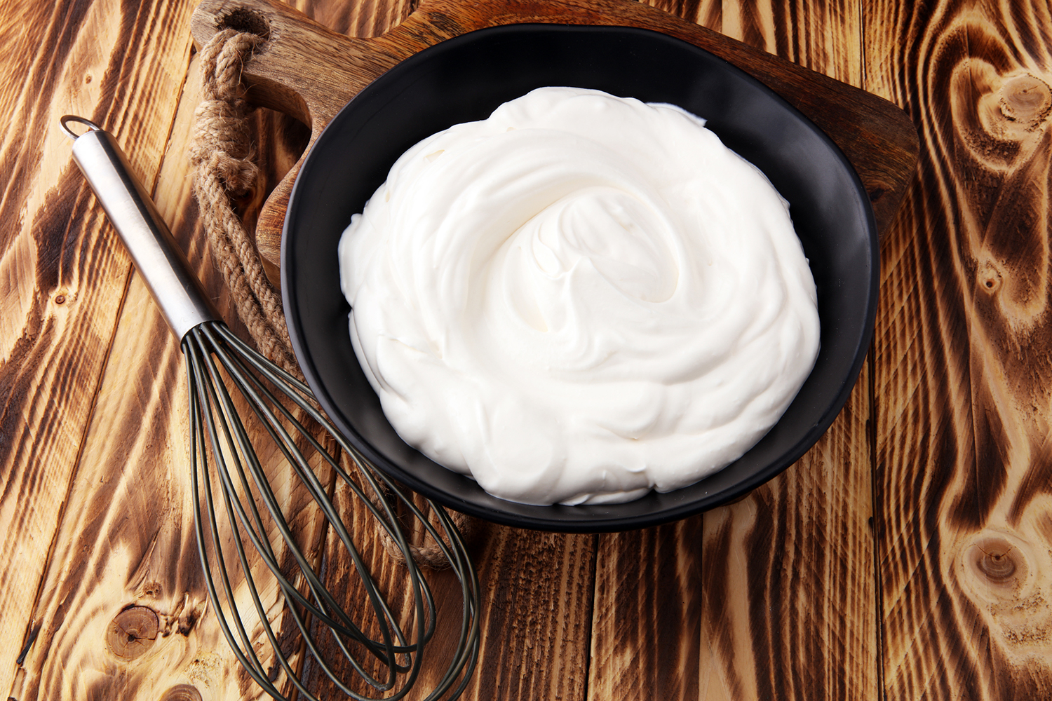 Homemade Whipped Cream in a black bowl with a whisk on a wooden table.