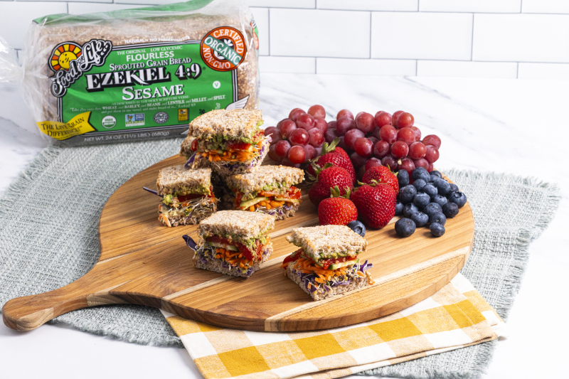 Colorful veggie sandwich on a cutting board, garnished with berries and grapes, with a package of Food for Life Ezekiel 4:9 bread in the background