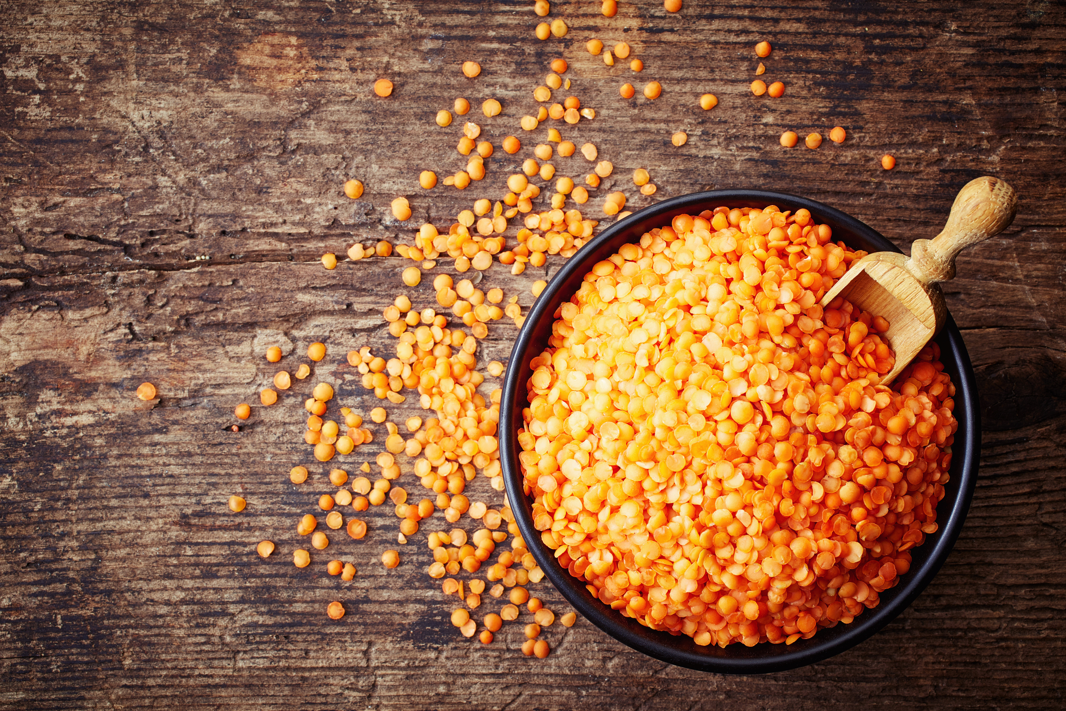 A bowl of uncooked red lentils with a wooden scoop in it. 