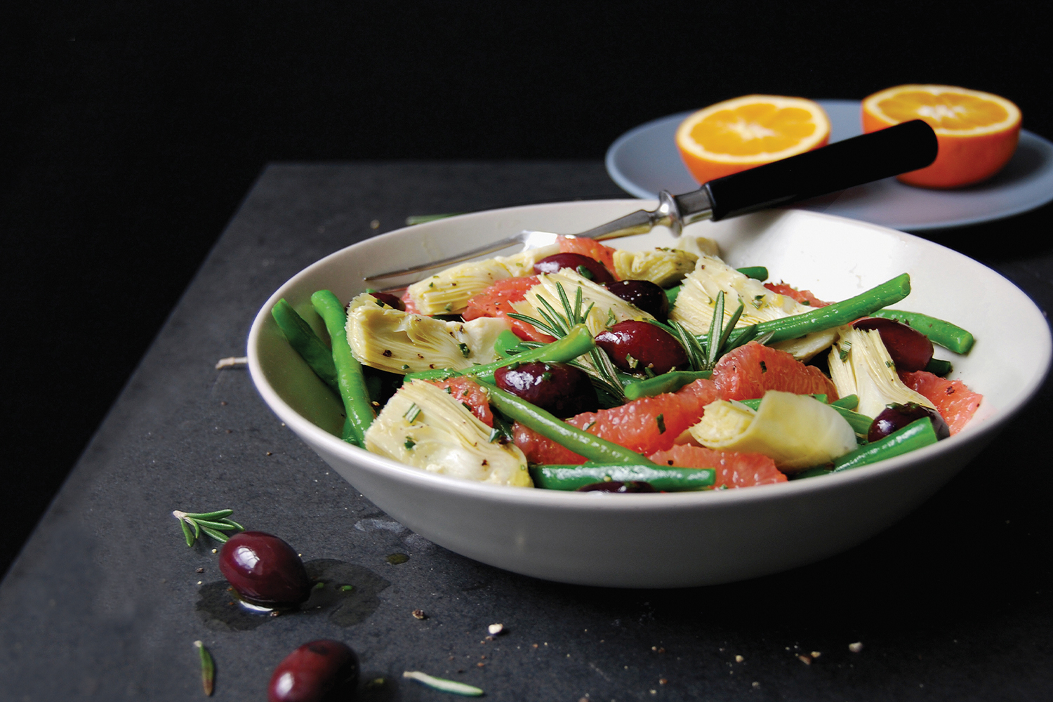 Green Bean, Artichoke, & Grapefruit Salad with Olives and Rosemary in a white bowl with a cut orange in the background. Black background.