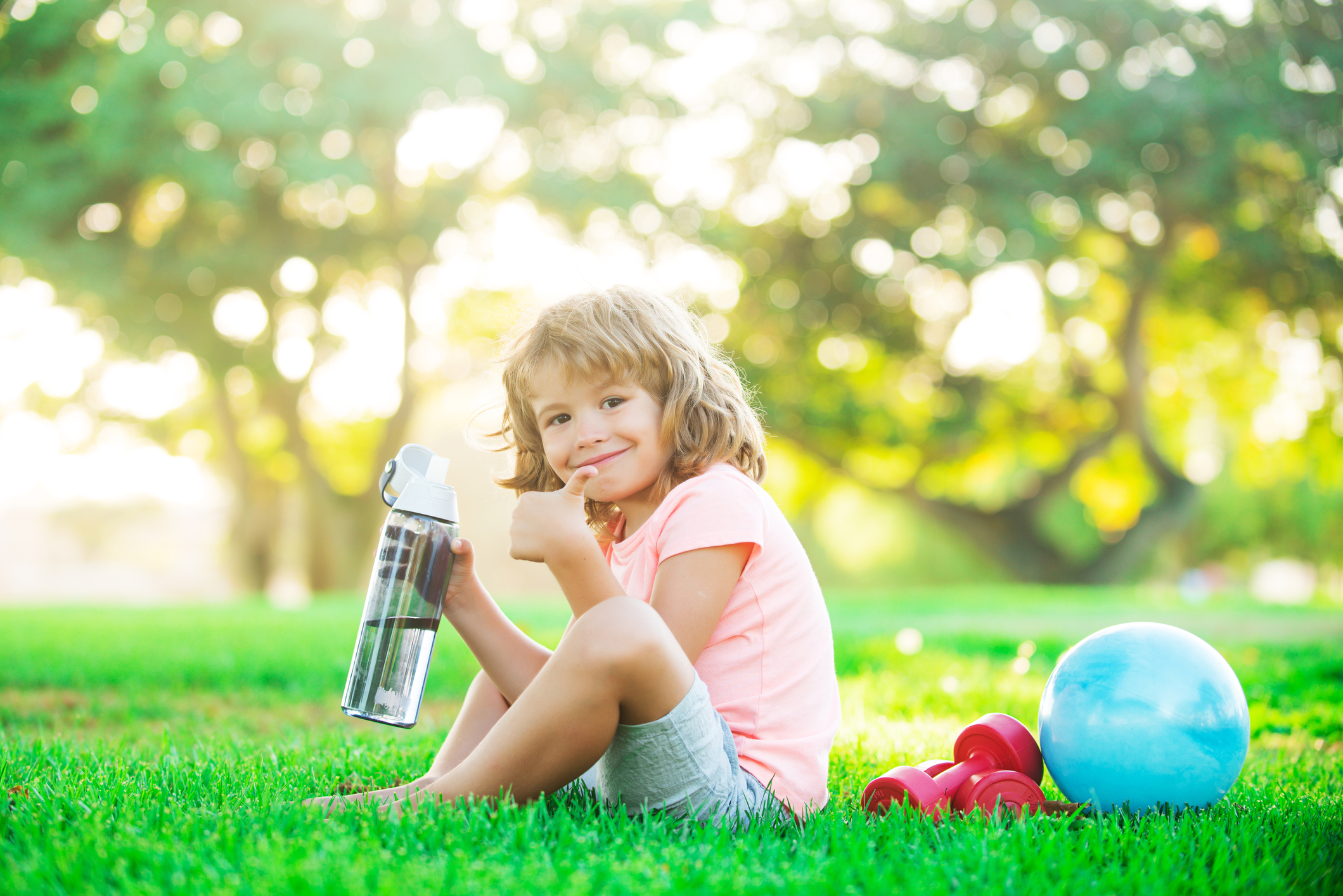 Kid with a water bottle sitting on the grass outside. 