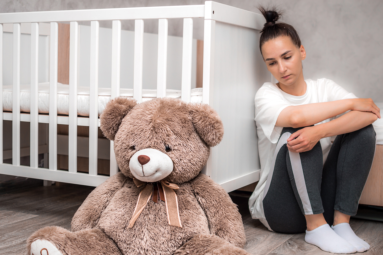 a depressed woman leaning on a crib looking at a teddy bear
